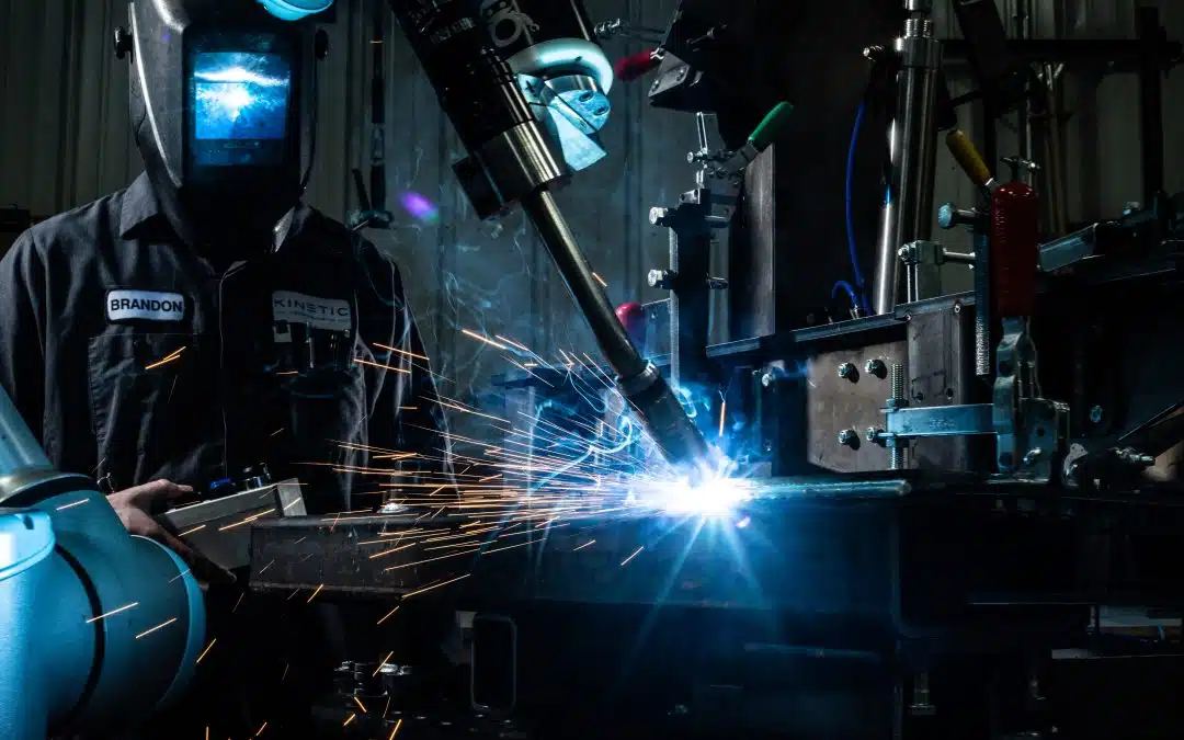 Welder operating a robotic arm for precision welding, with sparks flying in a workshop